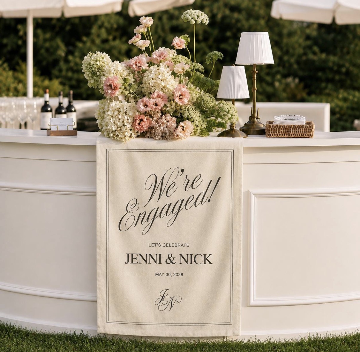 Linen We're Engaged sign hanging on a white bar with florals and lamps